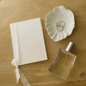 May include: A white booklet with the word "VOWS" embossed on the cover, a white ceramic dish with two rings, and a clear glass bottle of perfume on a wooden surface.