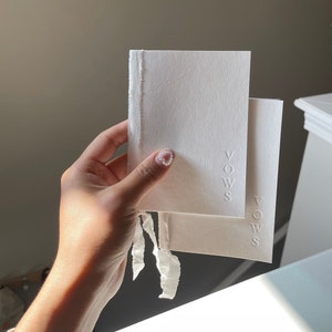 May include: Two white cards with the word "VOWS" embossed on them. The cards are tied together with a white ribbon.