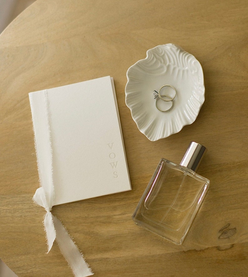 May include: A white booklet with the word "VOWS" embossed on the cover, a white ceramic dish with two rings, and a clear glass bottle of perfume on a wooden surface.