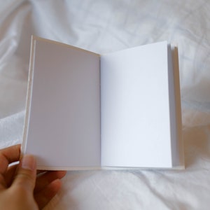 May include: A blank white notebook with a white cover and white pages is open and lying flat on a white bedspread.