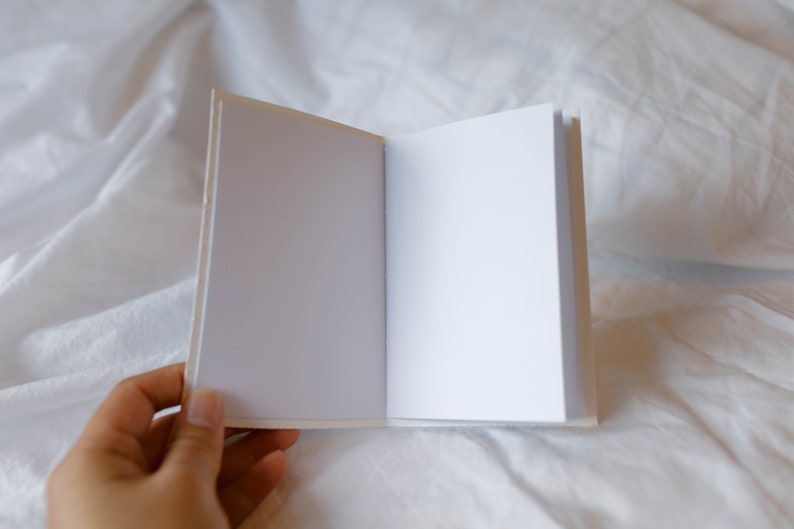 May include: A blank white notebook with a white cover and white pages is open and lying flat on a white bedspread.