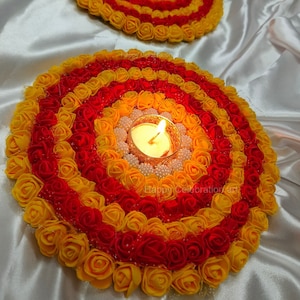 Könnte beinhalten: Drei kreisförmige Blumenarrangements mit roten und gelben Stoffrosen, die eine kleine Teelichtkerze in der Mitte umgeben. Der Text "Happy Celebration Art" ist auf zwei der Arrangements sichtbar.