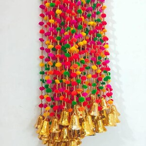May include: A colourful garland of pom-poms in red, green, yellow, and pink with gold bells hanging from the bottom.