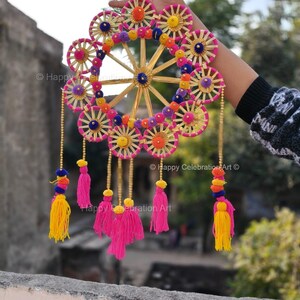Op de afbeelding: Een kleurrijke, handgemaakte wanddecoratie met een cirkelvormig ontwerp met roze, gele en oranje accenten. Het stuk bevat kwasten en kralenkettingen, waardoor een levendig en decoratief item ontstaat. Het ontwerp bevat een wielachtige structuur met spaken.