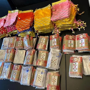 Op de afbeelding: Een verzameling kleurrijke stoffen zakjes in roze, oranje, geel en blauw, met gouden accenten en kwastjes. Daaronder liggen verschillende verpakkingen met decoratieve gezichtsjuwelen in diverse kleuren en ontwerpen.