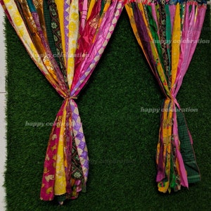May include: Two colorful fabric curtains made of strips of different fabrics, hanging in front of a green artificial grass backdrop. The curtains are tied together at the top and hang down to the bottom of the image.