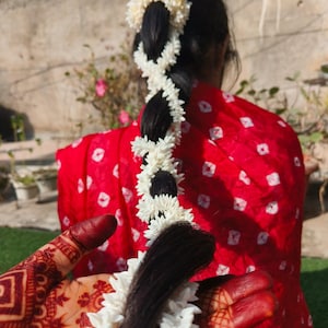 Può includere: I lunghi capelli neri di una donna sono intrecciati e decorati con fiori bianchi. I capelli sono tenuti in posizione da una ghirlanda di fiori bianchi. La donna indossa un vestito rosso con pois bianchi.