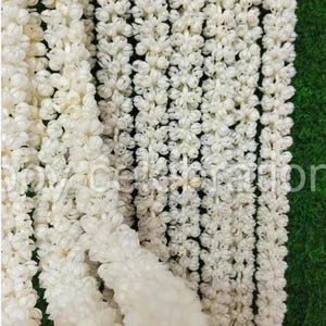 Op de afbeelding: Meerdere lange, verticale strengen van kleine, witte jasmijnbloemen die in een dicht, doorlopend patroon aan elkaar zijn geregen. De delicate, crèmekleurige bloesems zitten strak langs elk koord, wat een uniforme, gestructureerde bloemenslinger vormt.