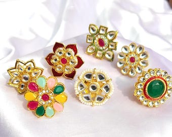 May include: Six gold-tone rings with intricate designs featuring colorful gemstones and mirrored accents. The rings are arranged on a white fabric background.