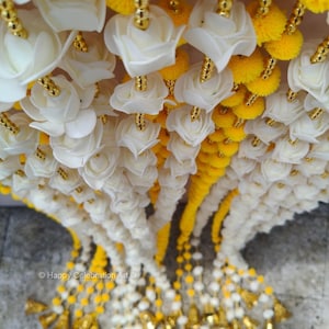 May include: Overhead view of hanging garlands with white rose-shaped flowers, yellow pom-poms, and gold detailing. The garlands create a layered effect. The text "Happy Celebration Art" is visible.