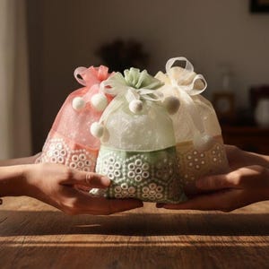 May include: Three decorative organza bags in shades of pink, green, and cream, each tied with a ribbon and adorned with small white pom-poms. The bags have a decorative, openwork design at the base. The bags are being held by two hands.