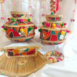 May include: Two colorful, decorated, round, fabric pots with gold trim and tassels. The pots are on a white surface with a woven bamboo tray and a small, colorful, fabric ring.