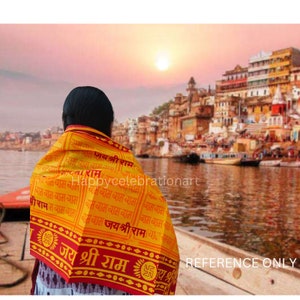 Può includere: Uno scialle giallo e rosso con il testo "Jai Shri Ram" stampato su di esso. Lo scialle è drappeggiato sulle spalle di una persona. La persona è in piedi su una barca e guarda un paesaggio urbano sullo sfondo. Il paesaggio urbano è composto da edifici con muri arancioni e gialli. Il cielo è di un colore rosa e arancione tenue. Il sole sta tramontando in lontananza.