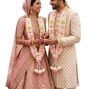 Puede incluir: Un lehenga rosa empolvado con intrincados bordados dorados, junto a un dupatta y una guirnalda floral a juego. Al lado, un sherwani color crema con patrones dorados, turbante a juego y guirnalda floral, ambos con detalles de flores en capas.