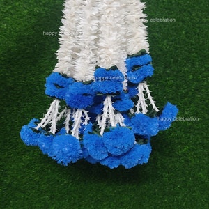 Puede incluir: Una guirnalda de flores artificiales blancas y azules. La guirnalda está hecha de flores blancas con detalles azules. La guirnalda está colgada de una mano. El texto "happy celebration" es visible en la guirnalda.