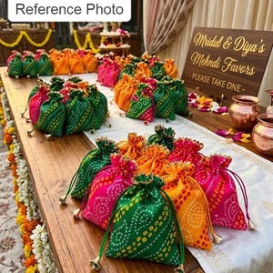 May include: A collection of small drawstring pouches in vibrant green, orange, and pink with a white dotted pattern. A wooden sign reads "Mridul & Diya's Mehndi Favors -PLEASE TAKE ONE-". The pouches feature metallic bell-shaped cord ends.