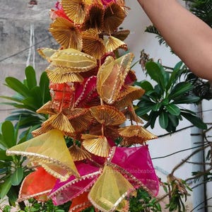 May include: A string of decorative hanging ornaments made of gold, pink, orange, and yellow fabric. The ornaments are arranged in a cascading pattern and have a fringed edge.