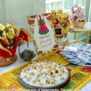 May include: A table display with various items for a celebration. It includes a sign that reads "Mehadi Bazaar Please pick one of each", jewelry, and decorative items in gold, red, and white. A floral arrangement is also present.