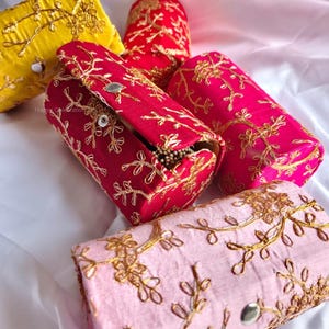 Puede incluir: Cuatro pequeñas cajas cilíndricas con diseños florales bordados. Las cajas son en tonos de rosa, amarillo y rojo con bordados dorados. Las cajas son de tela y tienen un cierre de botón.