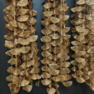 May include: Three strands of gold-colored paper garland, each strand is made of small, fan-shaped paper pieces. The garlands are hanging against a dark background.