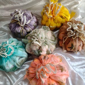 May include: An assortment of six decorative pouches in various colors, including yellow, purple, and peach. Each pouch is adorned with delicate pearl accents and tied with a ribbon. The pouches are filled with flowers and are arranged on a white satin surface.