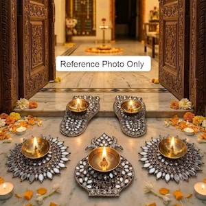 May include: Five brass diya oil lamps with lit wicks, arranged on a marble floor. Two lamps are shaped like feet, three are circular with mirrored petal accents. Scattered yellow and white marigold flowers surround the lamps near a doorway.
