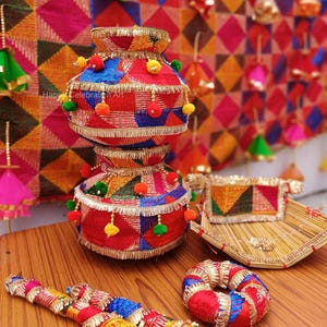 May include: Two decorative, stacked, red and blue embroidered pots with gold trim and colourful pom-poms. A woven tray and a decorative ring are also visible. The background features a colourful geometric pattern.