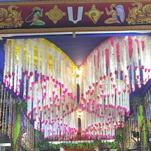 Puede incluir: Guirnaldas de flores blancas y rosas colgando de un techo, creando un arco decorativo. Las guirnaldas están hechas de flores artificiales y están enhebradas con hilo blanco. Las guirnaldas cuelgan de un techo azul y están iluminadas por luces de techo.