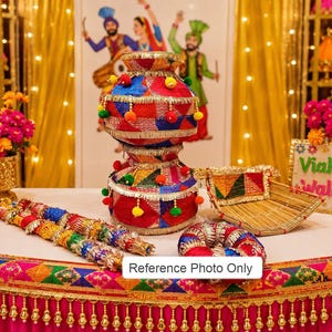 May include: A decorative set featuring a stacked, multi-colored fabric pot with pom-poms and gold trim, two matching fabric-wrapped sticks, a circular fabric ring, and a small woven tray, all displayed on a table with a pink, gold-trimmed cloth.