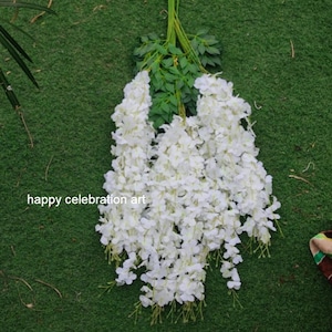 Puede incluir: Un ramo de flores de glicinia artificiales blancas con tallos y hojas verdes. Las flores están dispuestas en cascada, creando una exhibición exuberante y elegante.