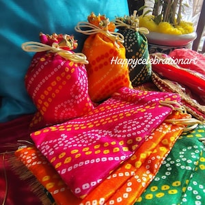 Może przedstawiać: Stos kolorowych materiałowych torebek z białym i żółtym wzorem. Torebki są związane złotym sznurkiem i mają złotą frędzlę. Torebki leżą na czerwonej powierzchni.