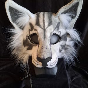 Peut inclure: Un masque félin blanc et gris avec des oreilles pointues, de longues touffes de fourrure blanche sur les joues, des marques gris foncé autour des yeux et sur le front, et un nez noir. Le masque présente de grandes ouvertures oculaires ovales.