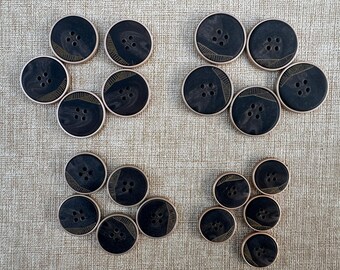 May include: A collection of dark brown buttons with four holes, arranged in a floral pattern. Each button has a decorative gold-colored design and a light-colored rim. The buttons are on a textured, neutral-colored surface.