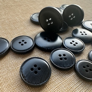 May include: A collection of black buttons with four holes each, in various sizes. The buttons are arranged on a textured, tan surface. The buttons have a glossy finish and are suitable for sewing or craft projects.