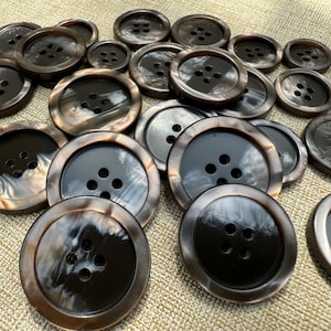 May include: A selection of brown and black buttons, each with four holes. The buttons have a glossy, marbled effect and are of varying sizes. They are arranged on a textured, beige surface.