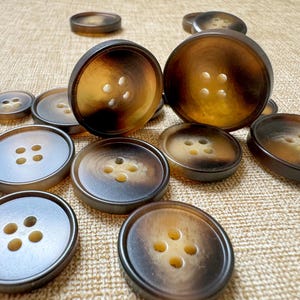 Puede incluir: Un conjunto de botones marrones y beige con cuatro agujeros cada uno. Los botones, de varios tamaños, están dispuestos sobre una superficie beige texturizada. Los botones tienen un acabado brillante y un patrón de color degradado.