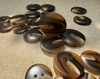 May include: A collection of large, oval-shaped buttons in shades of brown, tan, and black. The buttons have a marbled appearance with two or four holes for sewing. The buttons are scattered on a textured, tan surface.