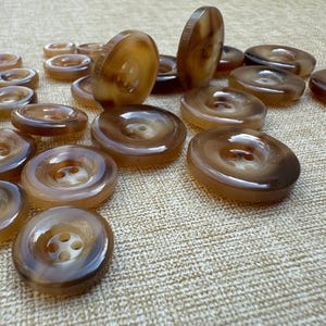 May include: A selection of brown and tan buttons with four holes. The buttons are of various sizes and made from a glossy, swirled material. Some buttons are standing upright, showing their thickness and design. These buttons are suitable for sewing and craft projects.