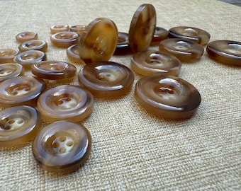 May include: A collection of brown and tan buttons with four holes. The buttons vary in size and are made of a glossy, swirled material. Some buttons are standing upright, showcasing their thickness and design. These buttons are suitable for sewing and craft projects.