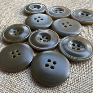 Op de afbeelding: Een verzameling olijfgroene knopen met elk vier gaatjes. De knopen hebben een gladde, matte afwerking en zijn gerangschikt op een getextureerde, beige stoffen achtergrond.