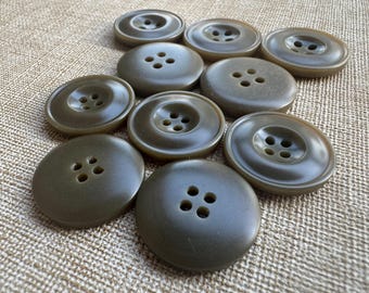 以下が含まれることがあります： オリーブグリーンのボタンのコレクション。各ボタンには4つの穴があり、滑らかでマットな仕上がりです。サイズと形状がわずかに異なり、ベージュの織り目加工の背景に配置されています。