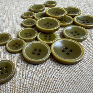 Può includere: Una collezione di bottoni rotondi color verde oliva di varie dimensioni, ciascuno dotato di quattro fori centrali e una superficie liscia leggermente incavata con un dettaglio del bordo circolare verde più chiaro, sparsi nel riquadro.