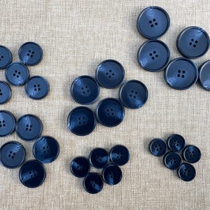 Puede incluir: Una colección de botones azul marino de varios tamaños, cada uno con cuatro agujeros. Los botones están dispuestos en varios grupos sobre un fondo de tela de tono neutro. Los botones tienen un acabado brillante.