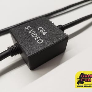 以下が含まれることがあります： 白文字で「S-VIDEO C64」と書かれた、C64コンソール用の黒いS-VIDEOアダプター。アダプターには2本の黒いケーブルが接続されています。画像には「GAMING FUN 80」というテキストが入った黄色のロゴが含まれています。