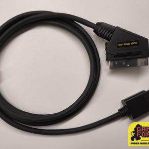 Puede incluir: Cable negro N64 RGB Mod con un cierre rojo. El cable tiene un conector SCART y un conector Nintendo 64. El texto "N64 RGB MOD" está impreso en el conector SCART. El logotipo de Gaming Fun 80 está en la esquina inferior derecha.