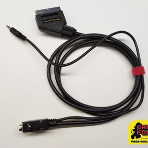 Peut inclure: Câble SCART noir avec une prise jack 3,5 mm et une attache de câble rouge. Le câble comporte un connecteur noir avec l'inscription "Analogizer VGA". L'image comprend un logo "Gaming Fun 80".