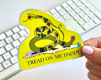 Könnte beinhalten: Ein gelber Aufkleber mit einer schwarzen und grünen Schlange mit einem roten Apfel im Maul. Die Schlange ist um ein schwarzes Band gewickelt. Der Aufkleber trägt den Text "Tread on me Daddy" in schwarzen Buchstaben.