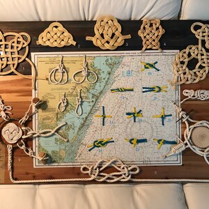 May include: A wooden board with a nautical chart in the center. The chart is surrounded by various rope knots, including a square knot, a figure eight knot, and a clove hitch. The knots are labeled with numbers. The board is a great way to learn about different types of knots.