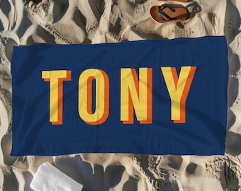 May include: A navy blue beach towel with the name "TONY" in large, yellow and orange block letters. The towel is rectangular and lies on a sandy beach, with a white towel, sandals, and a bow tie nearby.