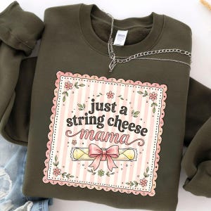May include: Olive green sweatshirt with a graphic that reads "just a string cheese mama." The graphic is a pink and white striped rectangle with a scalloped border, flowers, and a string cheese illustration with a pink bow.